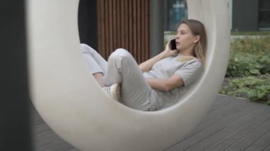 Genç bir kadın telefonda konuşuyor. Bahçede beyaz fütüristik bir salıncakta sallanıyor.