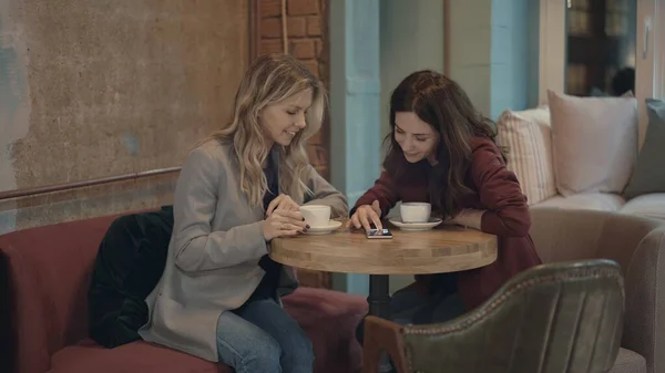 Dos mujeres atractivas en la cafetería charlando y discutiendo algunos chismes utilizando las redes sociales en el teléfono inteligente. El concepto de amistad y feminismo. — Foto de Stock