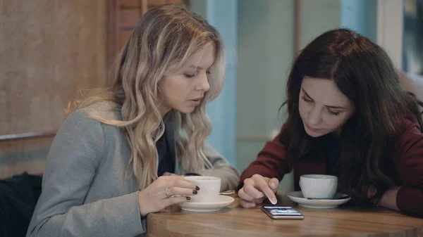 Dos mujeres atractivas en la cafetería charlando y discutiendo algunos chismes utilizando las redes sociales en el teléfono inteligente. El concepto de amistad y feminismo. — Foto de Stock