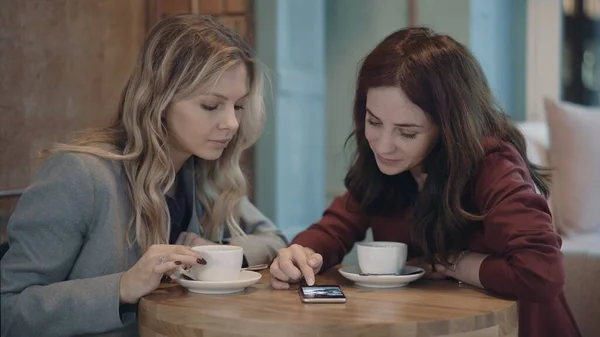 Dos mujeres atractivas en la cafetería charlando y discutiendo algunos chismes utilizando las redes sociales en el teléfono inteligente. El concepto de amistad y feminismo. — Foto de Stock