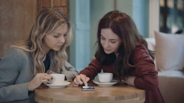 Kafede sohbet eden ve bazı dedikoduları tartışan iki çekici kadın akıllı telefonda sosyal ağları kullanıyor. Arkadaşlık ve feminizm kavramı. 