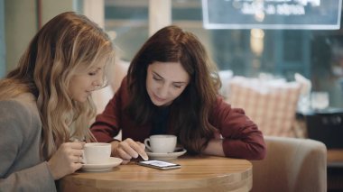 Kafede sohbet eden ve bazı dedikoduları tartışan iki çekici kadın akıllı telefonda sosyal ağları kullanıyor. Arkadaşlık ve feminizm kavramı. 