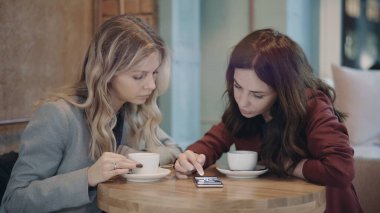Kafede sohbet eden ve bazı dedikoduları tartışan iki çekici kadın akıllı telefonda sosyal ağları kullanıyor. Arkadaşlık ve feminizm kavramı. 