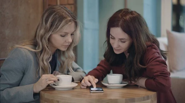 Dos mujeres atractivas en la cafetería charlando y discutiendo algunos chismes utilizando las redes sociales en el teléfono inteligente. El concepto de amistad y feminismo. — Foto de Stock