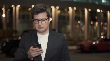 Gözlüklü bir iş adamı binanın arka planında telefonu kaydırıyor.