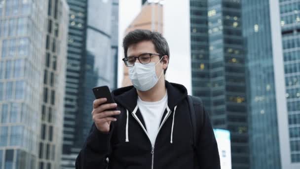 Άνδρας με προστατευτική μάσκα σε casual ρούχα κρατώντας ένα τηλέφωνο, φόντο του επιχειρηματικού κέντρου — Αρχείο Βίντεο