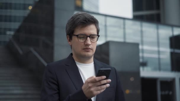 Мужчина в костюме стоит с телефоном на фоне стеклянных зданий — стоковое видео