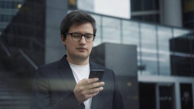 Takım elbiseli adam cam binaların arka planında telefonla ayakta duruyor.