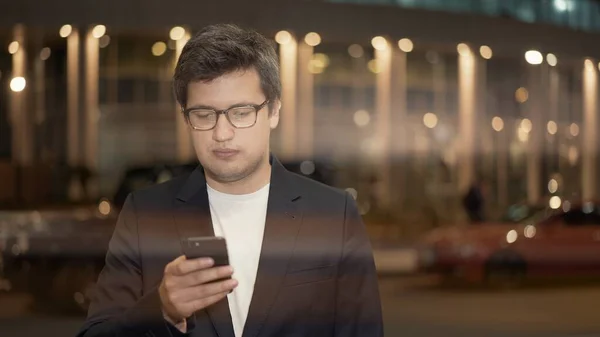Бізнесмен в окулярах прокручує телефон на фоні будівлі, блимає лінза — стокове фото