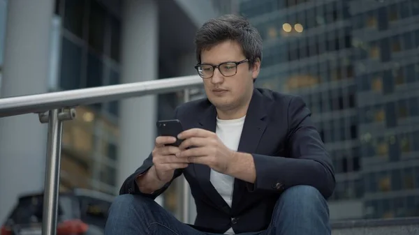 Бізнесмен сидить на сходах з телефоном на фоні скляних будівель, вечірній час — стокове фото