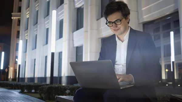 Man met laptop op schoot lachend op bank buiten de universiteit, lensflare — Stockfoto