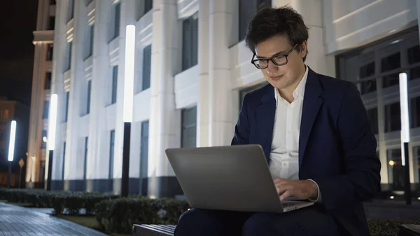 Man met laptop op schoot lachend op bank buiten de universiteit, laat werk — Stockfoto