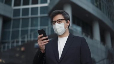İş adamı koruyucu bir maske takıyor ve iş merkezinin yakınında bir telefon tutuyor.
