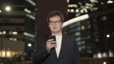 Gözlüklü bir iş adamı binaların ortasında telefonu kaydırıyor.
