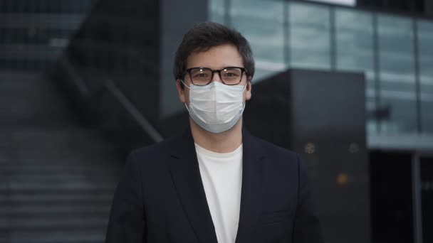 Geschäftsmann mit Maske blickt in die Kamera, Hintergrund von Geschäftsgebäuden — Stockvideo