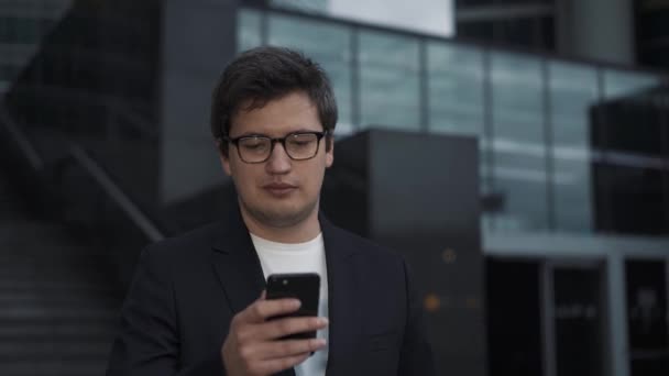 Мужчина в костюме стоит с телефоном на фоне стеклянных зданий — стоковое видео