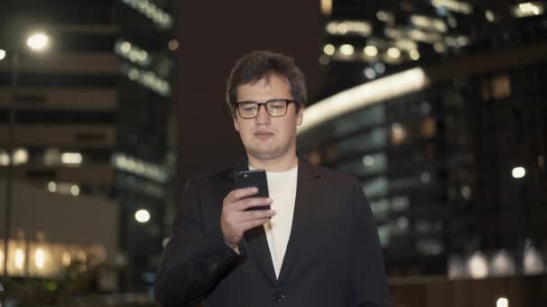 Бизнесмен в очках, прокручивающий телефон в центре зданий — стоковое видео