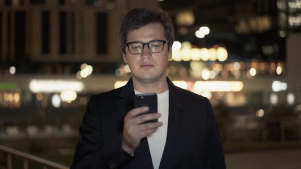 Geschäftsmann mit Brille scrollt das Telefon auf dem Hintergrund von Gebäuden — Stockvideo