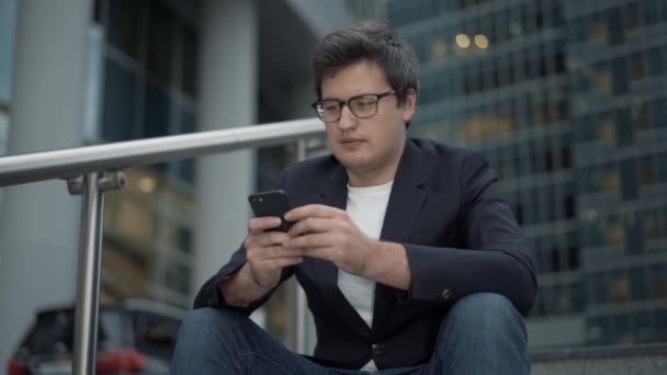 Бизнесмен, сидящий на лестнице с телефоном на фоне стеклянных зданий, вечернее время — стоковое видео
