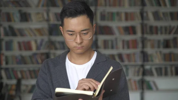 Asijský chlapec s brýlemi čtení knihy na pozadí knihoven — Stock fotografie