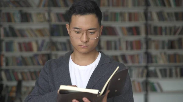 Asiatico ragazzo con occhiali in possesso di un libro su sfondo di librerie — Foto Stock