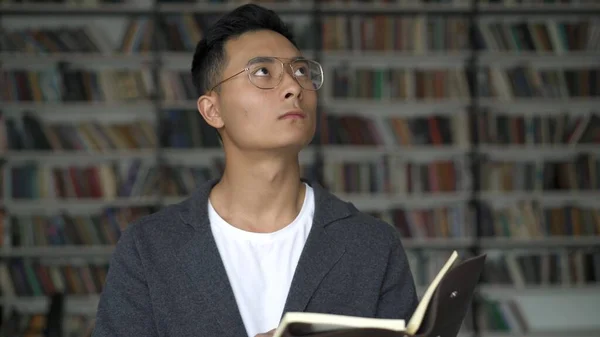Asian boy in eyeglasses holding a book with eyes up, background of bookshelves — Stock Photo, Image