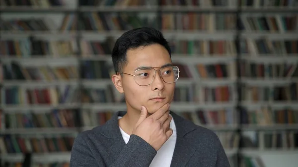 Estudante asiático em casaco cinza no fundo de estantes borradas na biblioteca — Fotografia de Stock