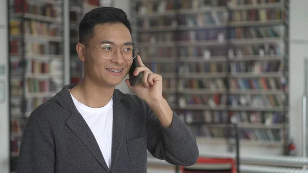 Ung leende asiatisk man talar i telefon, tänder hängslen, i biblioteket — Stockfoto