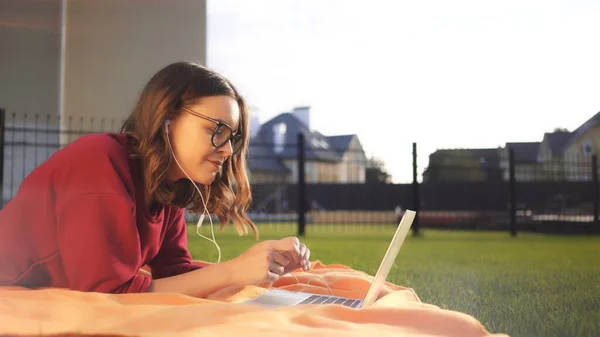 Gözlüklü ve dizüstü bilgisayarlı kız gülümsüyor çimlerde çalışıyor, güneşli akşam mercekleri parlıyor — Stok fotoğraf