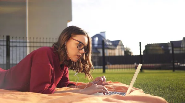 Chica en gafas graduadas y portátil que trabaja en el césped, resplandor de lente de noche soleado — Foto de Stock