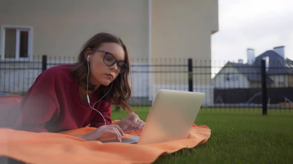 Jente i briller med øretelefoner og bærbar PC som arbeider på plenen – stockfoto