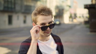 Genç adam kameraya bakıyor. Büyük gözlükler tutuyor. Flört ediyor.