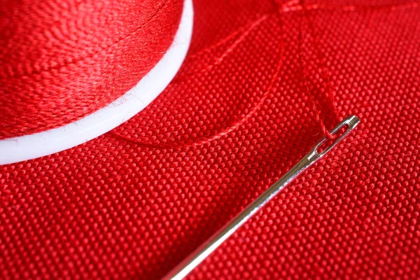 A close up image of a spool of red thread and sewing needle on red fabric.