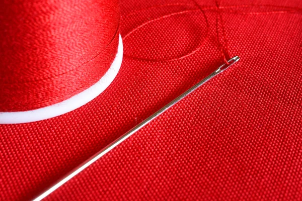 A close up image of a spool of red thread and sewing needle on red fabric.