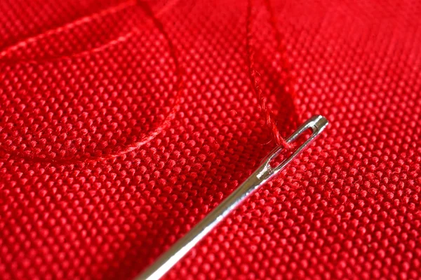 A close up image of a spool of red thread and sewing needle on red fabric.