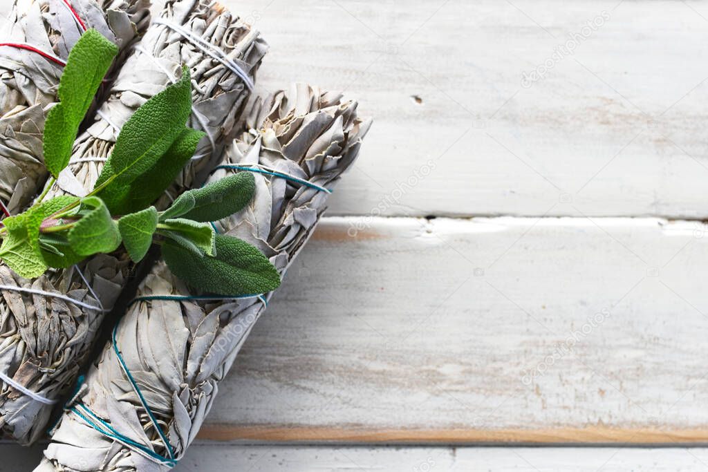 A top view image of white sage smudge bundles with fresh white sage leaves on a white wooden background. 