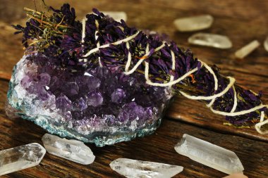 A close up image of an amethyst geode and lilac smudge stick with clear quartz crystals on a dark wooden desk top.  clipart