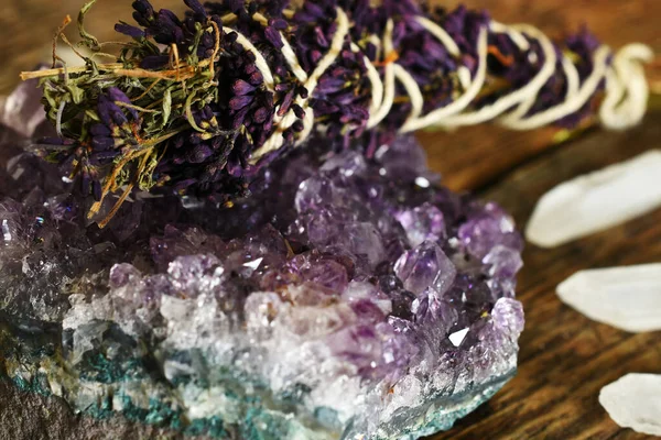 A close up image of an amethyst geode and lilac smudge stick with clear quartz crystals on a dark wooden desk top.