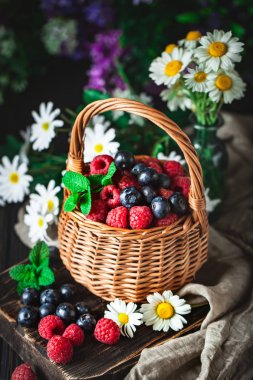 Ahududu ve yaban mersini bir sepetin içinde papatya çayı ve koyu arkaplanda yapraklar. Yaz ve sağlıklı gıda konsepti. Seçici odak.