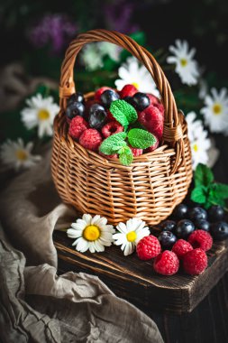 Ahududu ve yaban mersini bir sepetin içinde papatya çayı ve koyu arkaplanda yapraklar. Yaz ve sağlıklı gıda konsepti. Seçici odak.