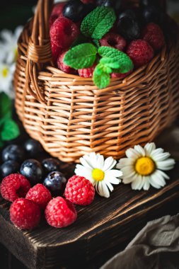 Ahududu ve yaban mersini bir sepetin içinde papatya çayı ve koyu arkaplanda yapraklar. Yaz ve sağlıklı gıda konsepti. Seçici odak.