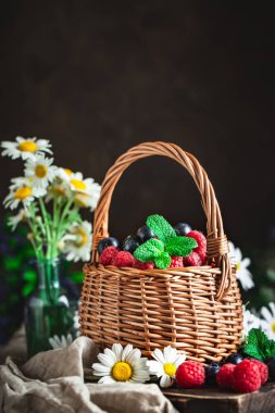 Ahududu ve yaban mersini bir sepetin içinde papatya çayı ve koyu arkaplanda yapraklar. Yaz ve sağlıklı gıda konsepti. Seçici odaklanma. Kopyalama alanı olan arkaplan.