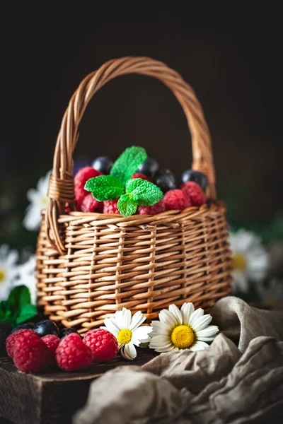 Frambuesas y arándanos en una cesta con manzanilla y hojas sobre un fondo oscuro. Verano y concepto de comida saludable. Enfoque selectivo . — Foto de Stock