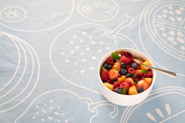 Mango, ahududu, kavun, yabanmersini ve çilekli beyaz kase mavi bir masa örtüsü üzerinde duruyor.