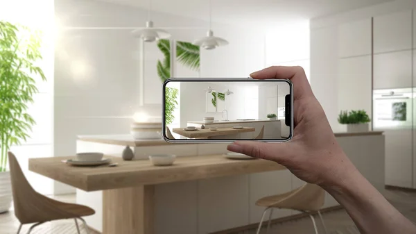 Hand holding smart phone, AR application, simulate furniture and interior design products in real home, architect designer concept, blur background, modern white and wooden kitchen