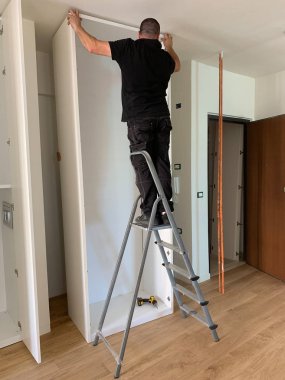 Marangozlar dolabın kapaklarını beyaz bir dolaba yerleştiriyor, dolapta yürüyorlar. Kısmen kurulu mobilyalar.