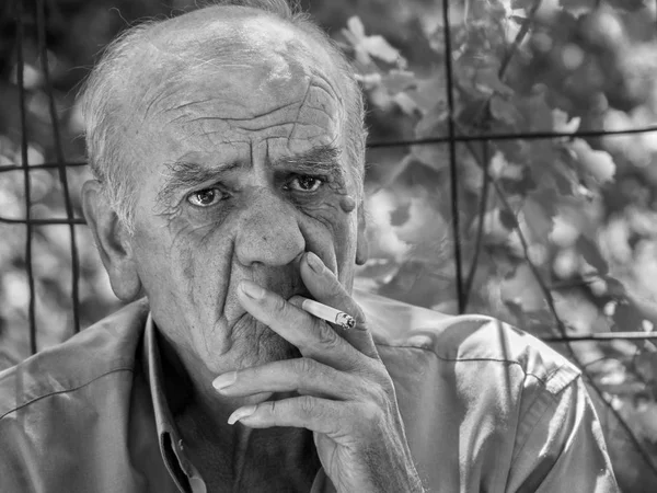 Closeup portrait of a serious old greek retired male who smokes a cigarette with a smile, in black and white