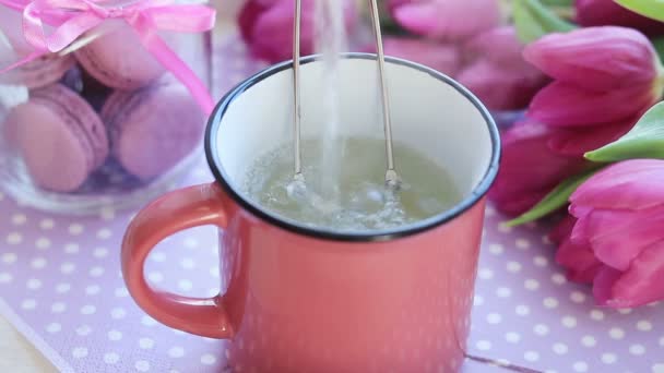 ピンクのセラミック カップにティーバッグに沸騰したお湯を注ぎ ピンクのチューリップと紅茶のカップのそばのテーブルの上のマカロン — ストック動画