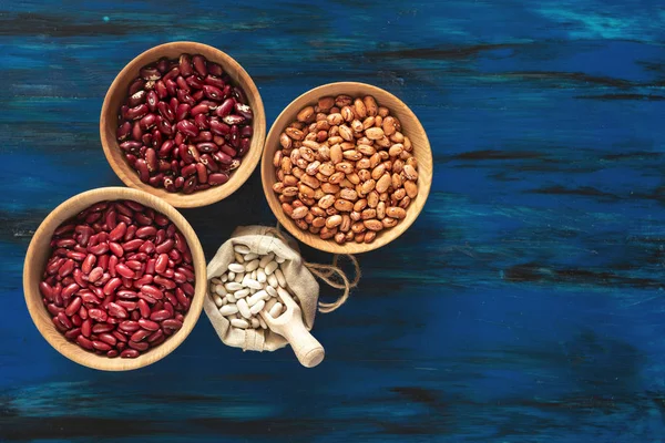 Assortment of kidney beans in wooden dishes and white kidney beans in linen bag on dark blue wooden background, mung bean, red kidney bean, white bean, brown bean.