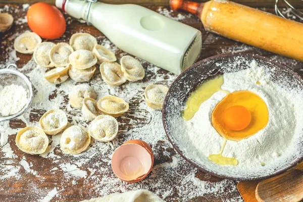 Ingredients and tools for baking - flour, eggs and glass of milk and raw homemade delicious traditional italian ravioli, dumplings on the wooden rustic table background, selective focus